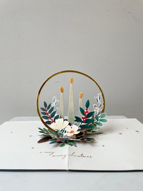 Interior of Pop up card laid on flat surface with gold candle ring and winter greenery on white background. Text says a very merry Christmas. 