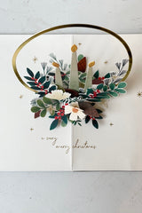 Top view of Interior of Pop up card laid on flat surface with gold candle ring and winter greenery on white background. Text says a very merry Christmas. 