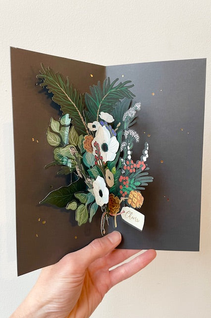 Hand holding pop up card open to display interior of bouquet of winter foliage and flowers with gift tag on black background. Text reads cheers.