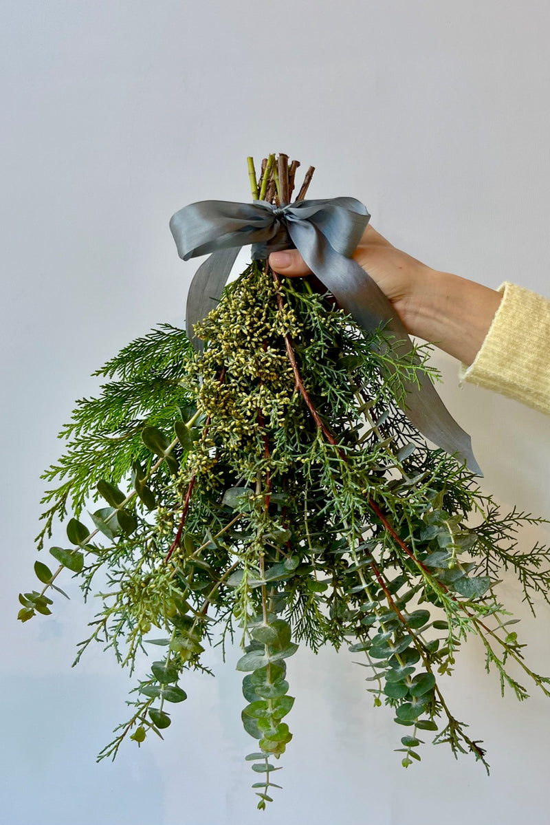 Hand holding a mixed greenery bouquet with eucalyptus, cypress, cedar tied with ribbon against a white wall. 