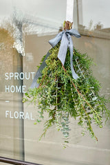 Hand holding a mixed greenery bouquet with eucalyptus, cypress, cedar tied with ribbon hanging on the glass door to the Sprout Home Floral store.  