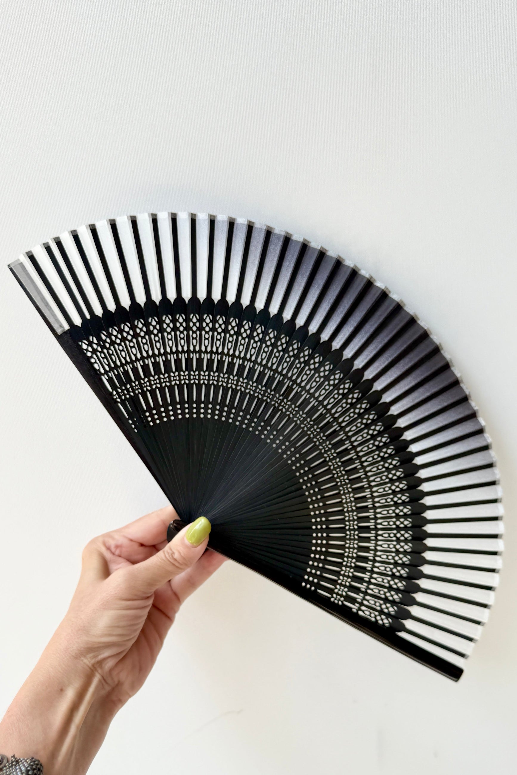 A hand holding a Silk Folding Fan White Silk w/ Black Bamboo with intricate white geometric patterns on the underside, set against a plain white background. ©Sprout Home