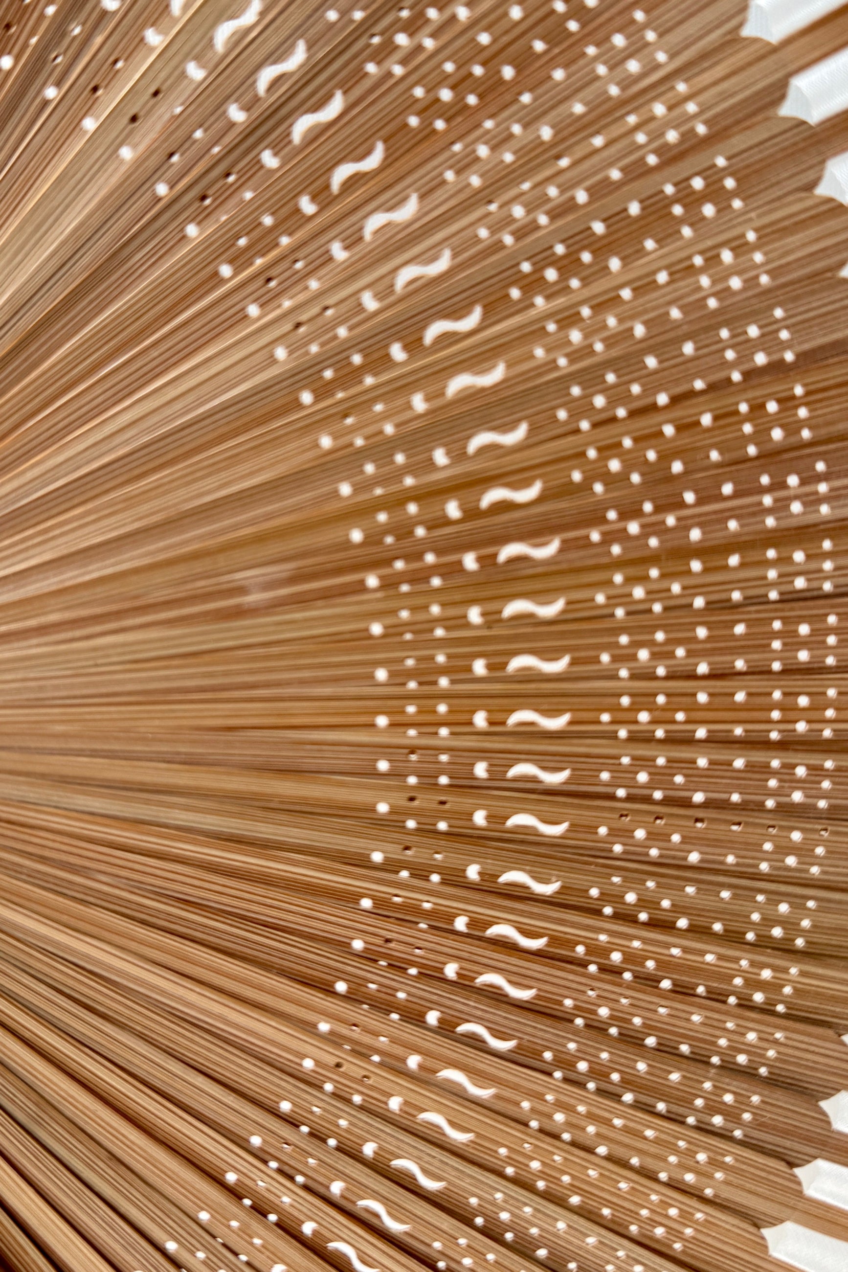 Close-up of a Silk Folding Fan Light Beige w/ Brown Bamboo made of many thin tan bamboo radiating from the center, with small white wave-like cutouts or holes arranged in rows along the right side. ©Sprout Home