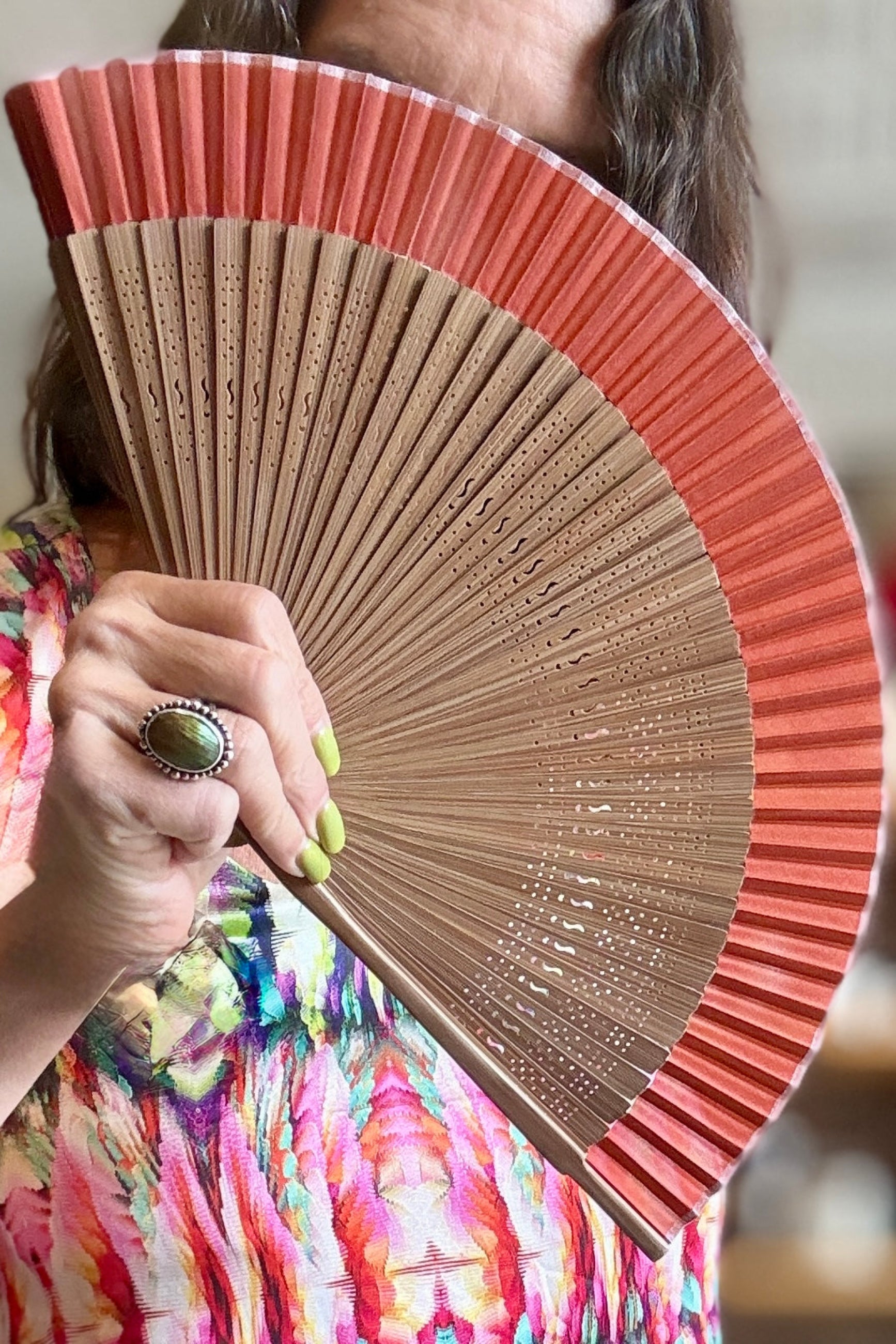 A close-up of a person holding a fSilk Folding Fan Harvest Brown w/ Brown Bamboo The fan has a warm coral-pink outer section and a natural light wood inner section. ©Sprout Home