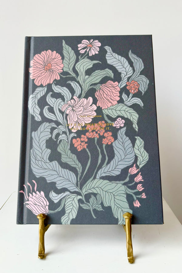 Hard cover notebook with pink and green floral motif against a black background with the phrase "Let's do what we love and do a lot of it" in gold lettering, displayed in a gold bookstand against white background