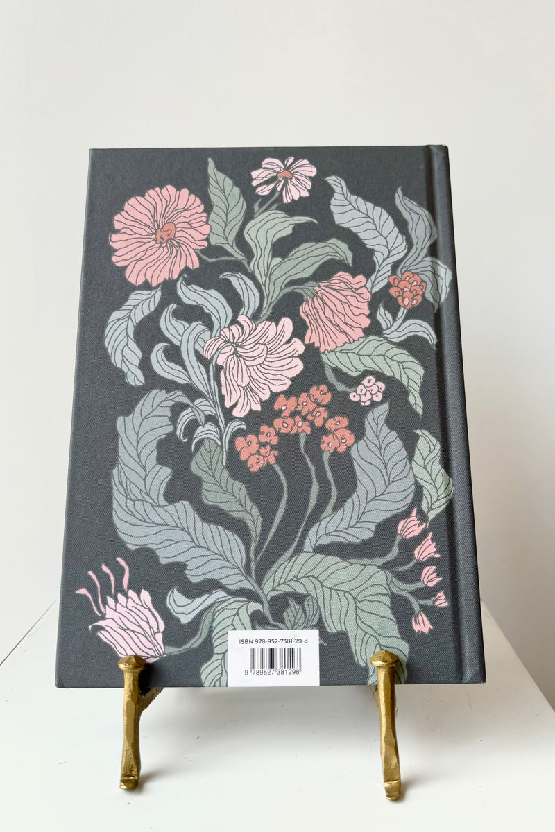 Back cover of notebook with pink and green floral motif against a black background displayed in a gold bookstand against white background