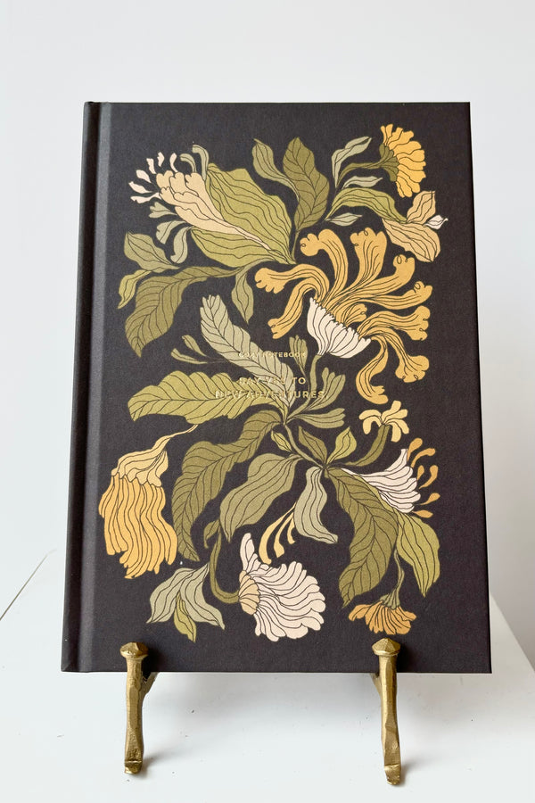 Hard cover notebook with gold, white and green floral motif against a black background with the phrase "Say yes to new adventures " in gold lettering, displayed in a gold bookstand against white background