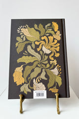 Back side of hard cover notebook with gold, white and green floral motif against a black background with the phrase "Say yes to new adventures " in gold lettering, displayed in a gold bookstand against white background