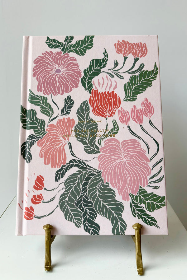 Hard cover notebook with pink, red and green  floral motif against a pink background with the phrase "You are exactly where you need to be" " in gold lettering, displayed in a gold bookstand against white background