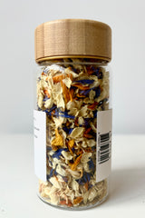 Back view of glass jar with dried flower petals composed of Jasmine, Tagete, and Cornflower petals with a woodgrain lid against white background