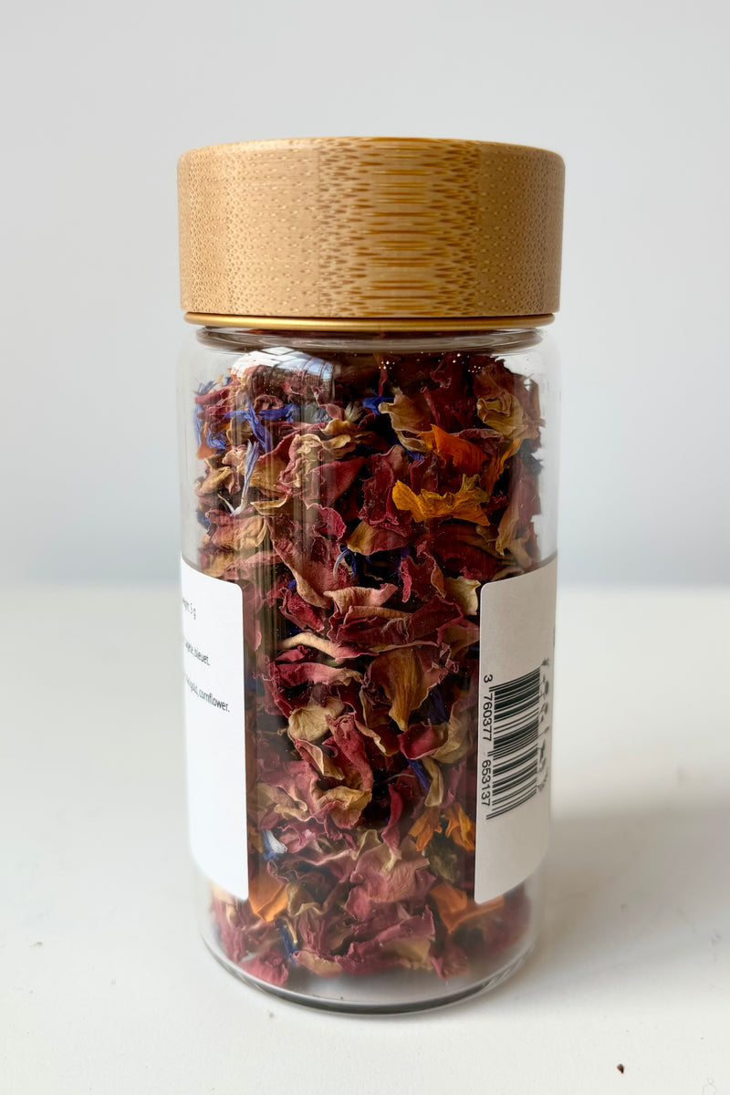 Back view of glass jar with dried flower petals composed of rose, marigold and cornflower petals against white background
