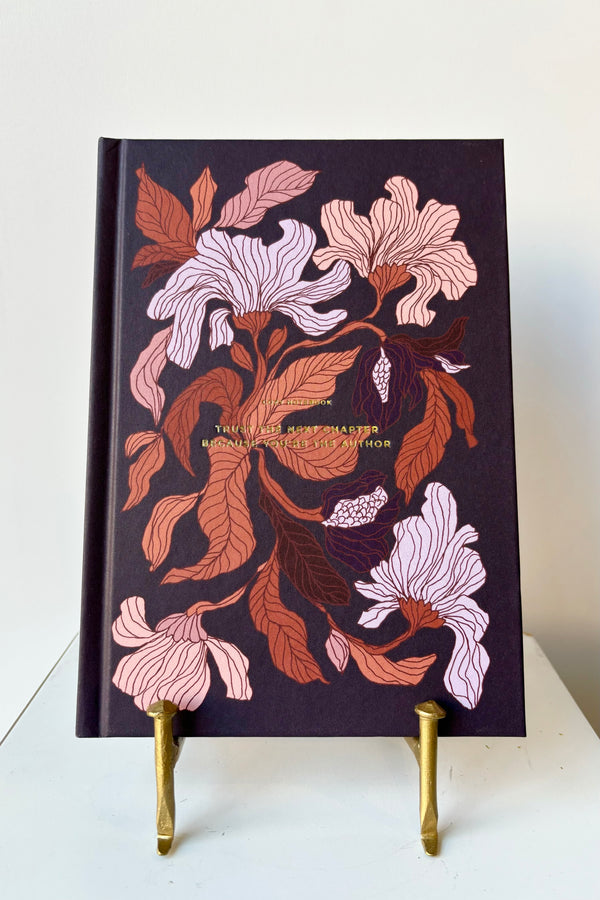 Hard cover notebook with a burgundy, peach and rust floral motif against a dark purple background with the phrase "Trust the next chapter because you're the author" in gold lettering, displayed in a gold bookstand against white background