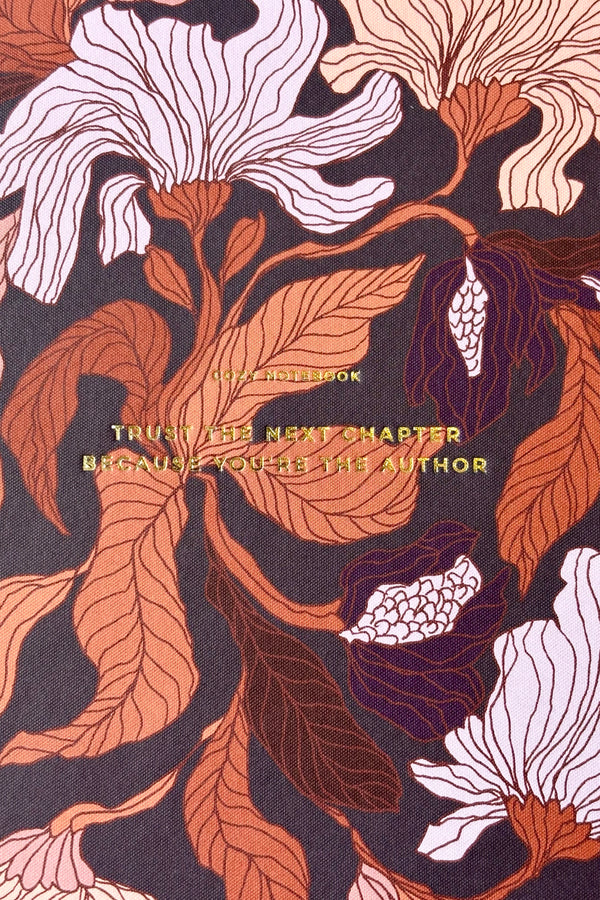 Close up of notebook with a burgundy, peach and rust floral motif against a dark purple background with the phrase "Trust the next chapter because you're the author" in gold lettering