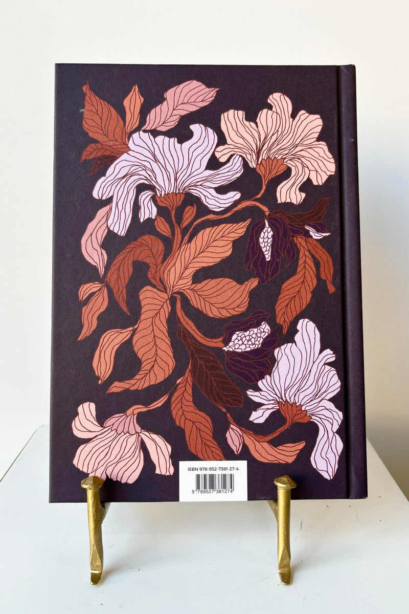 Back cover of notebook with a burgundy, peach and rust floral motif against a dark purple background with the phrase "Trust the next chapter because you're the author" in gold lettering, displayed in a gold bookstand against white background