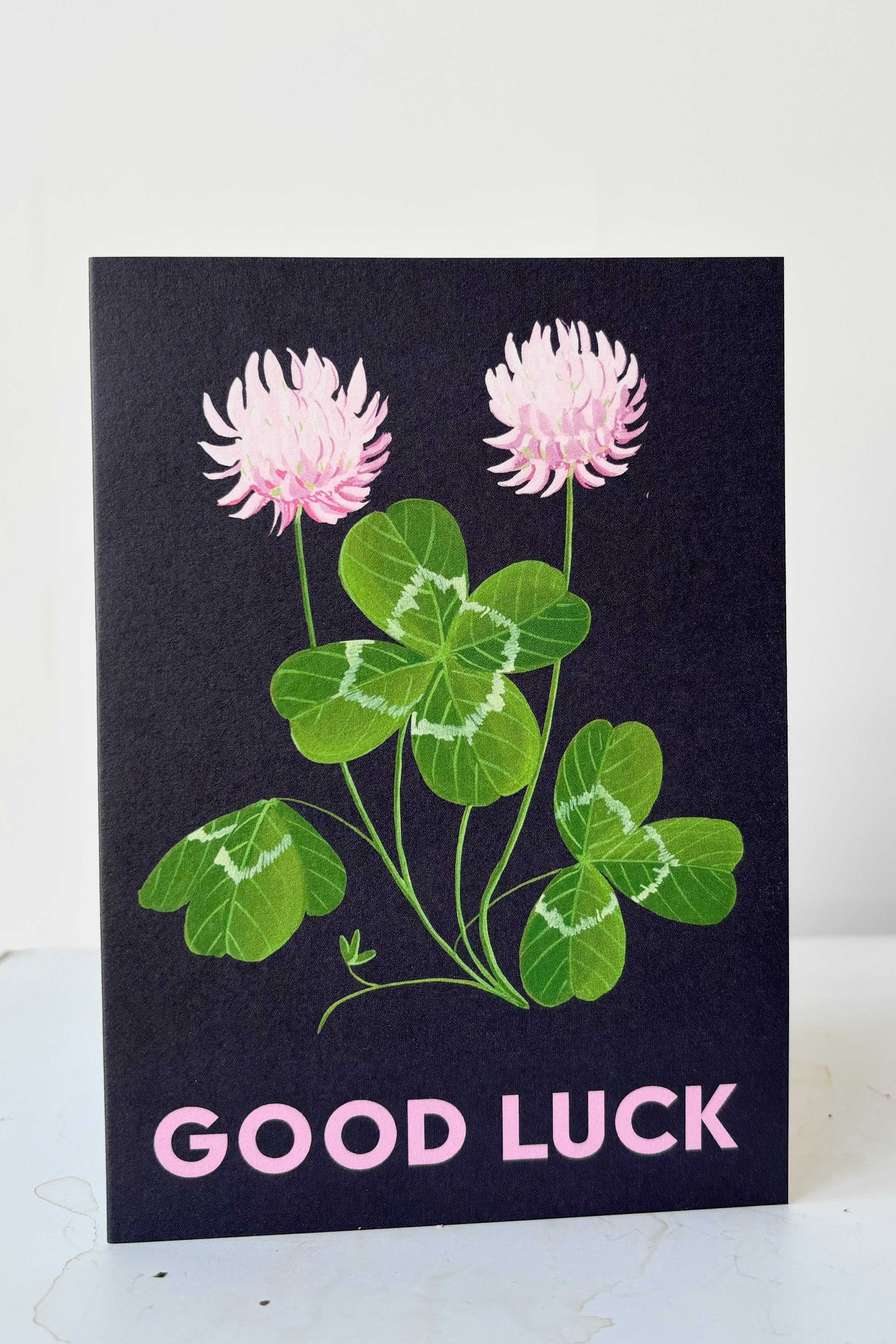 Clover greeting card with image of clover plant with pink flowers on dark blue background with the text  Good Luck on the front, blank inside against white background.