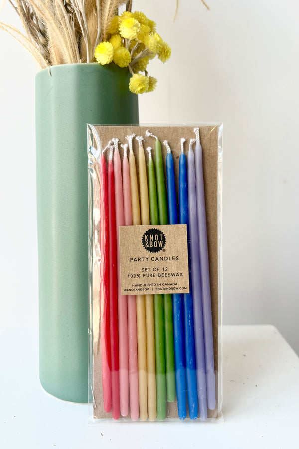 Image of twelve, thin, multicolored beeswax candles in red, pink, yellow, green, blue and purple in cellophane wrap against white bakcground