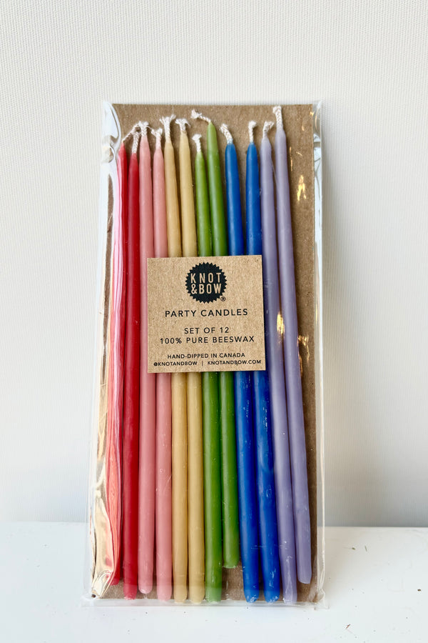 Close up image of twelve, thin, multicolored beeswax candles in red, pink, yellow, green, blue and purple in cellophane wrap against white bakcground