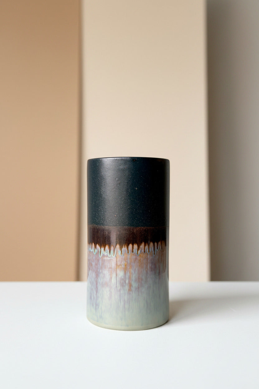 A handmade ceramic Cylinder Vase Z002 Extra Large with a dripping glaze finish, featuring a dark charcoal top half, a brown middle band, and a light blue-gray base on a white surface against a white wall. ©Sprout Home