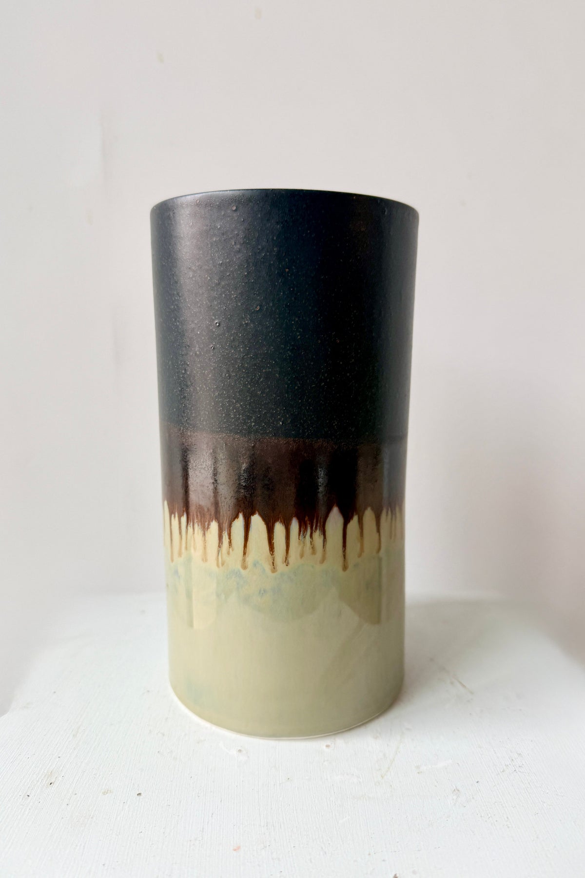 A handmade ceramic Cylinder Vase Z002 Extra Large dripping glaze design, featuring a matte black top half, a dark brown middle band, and a cream-colored bottom half. ©Sprout Home