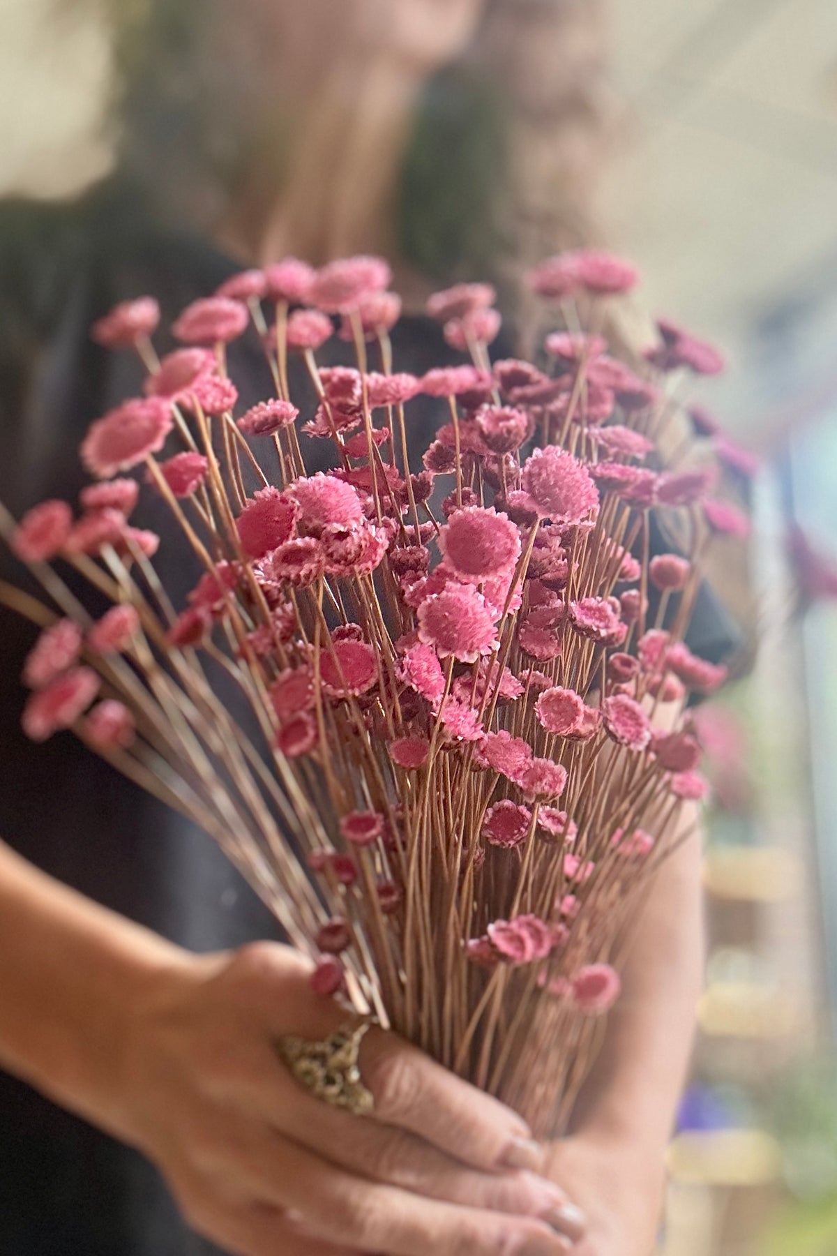 A person holding a Margaret Light Pink Color Preserved Bunch pink flowers with long brown stems, photographed in a softly blurred indoor setting. ©Sprout Home