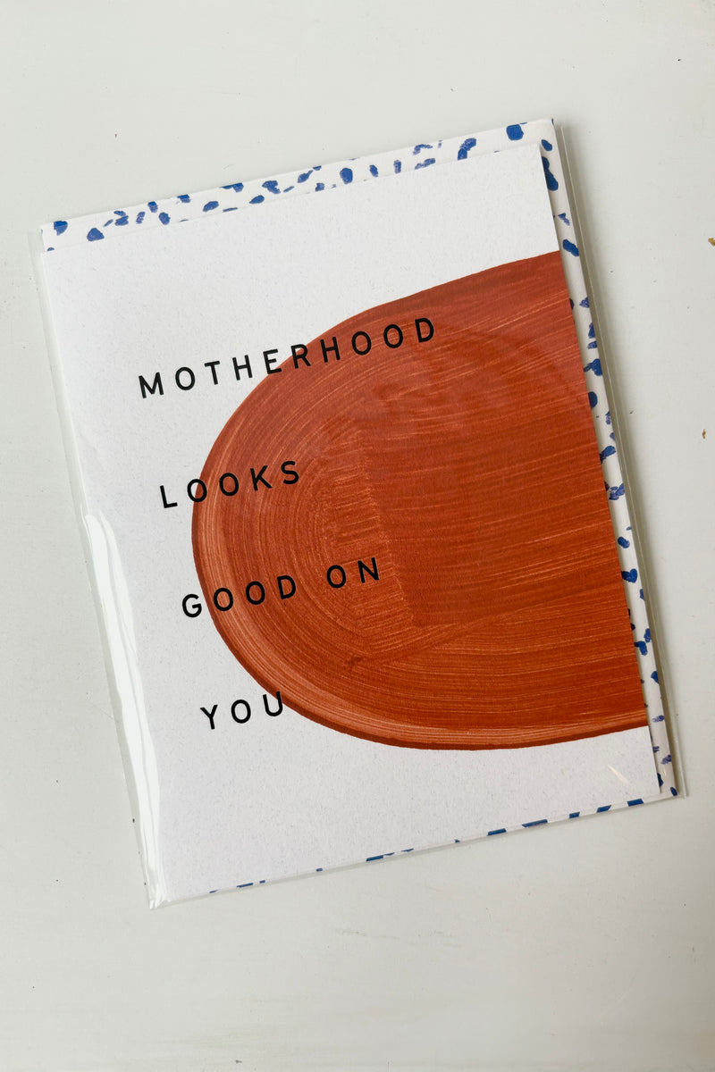 Painted greeting card with brown abstract design against white background with blue lettering that says motherhood looks good on you