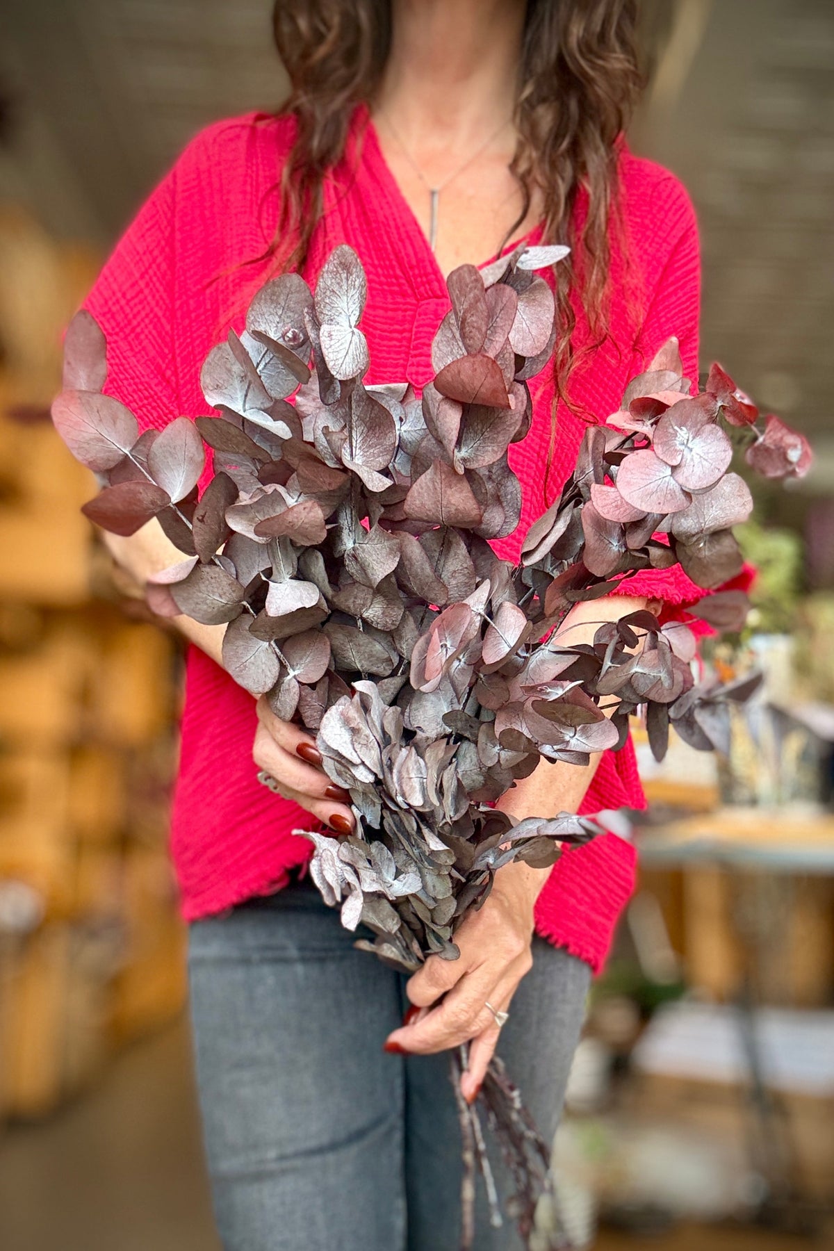 A person wearing a bright red shirt holds a Eucalyptus Spiral Burgundy Color Preserved Bunch in a shop setting. ©Sprout Home