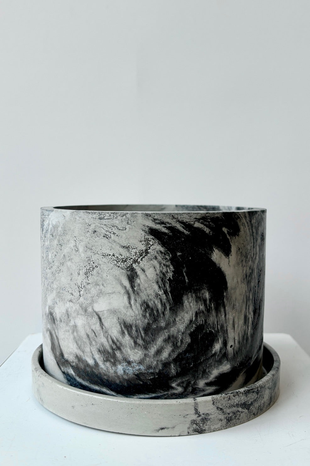 Marbled black and white varnished cement planter with drain hole and saucer against a white wall