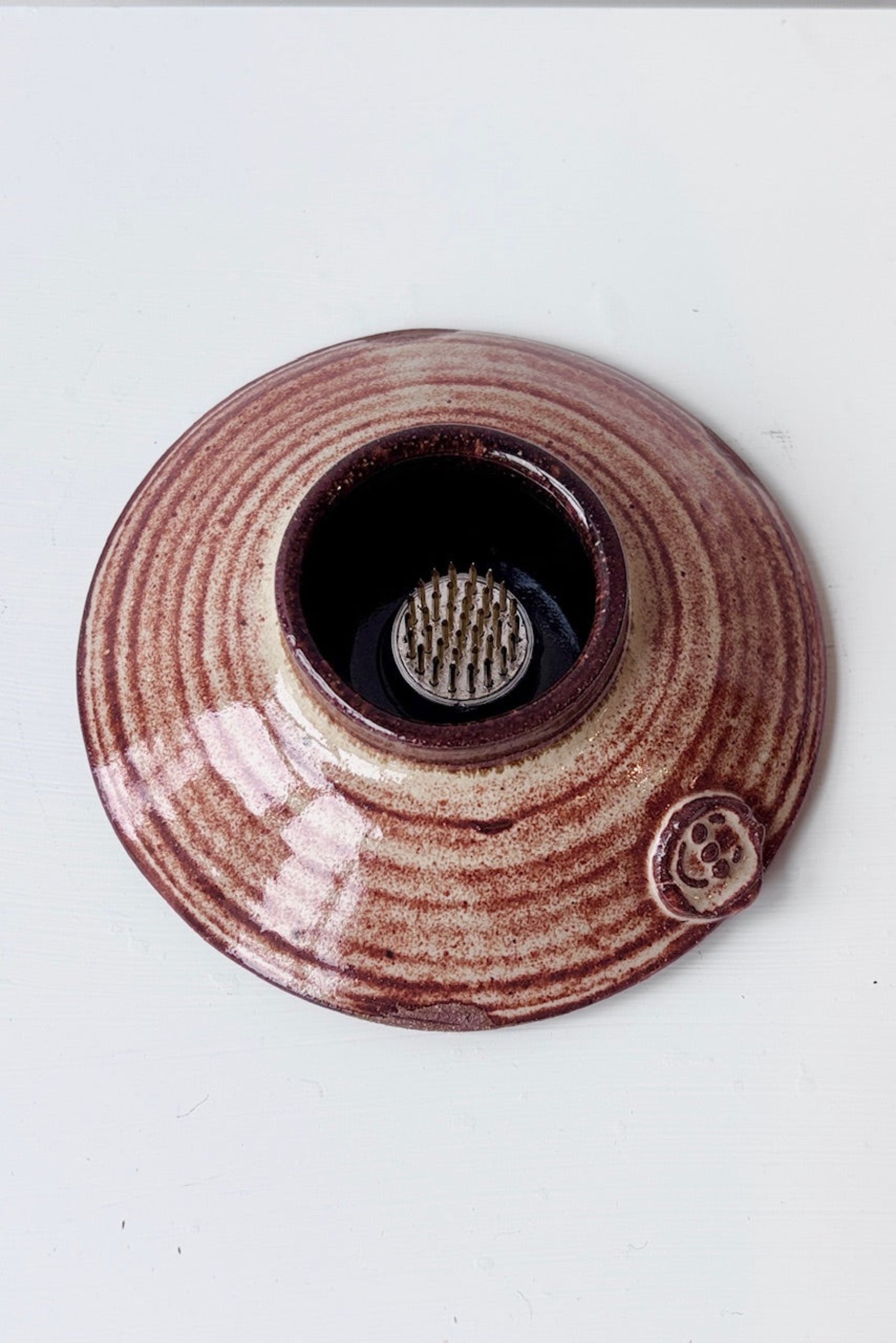 Overhead view of rust and cream colored, round, low Ikebana vase with attached floral frog against a white surface ©Sprout Home