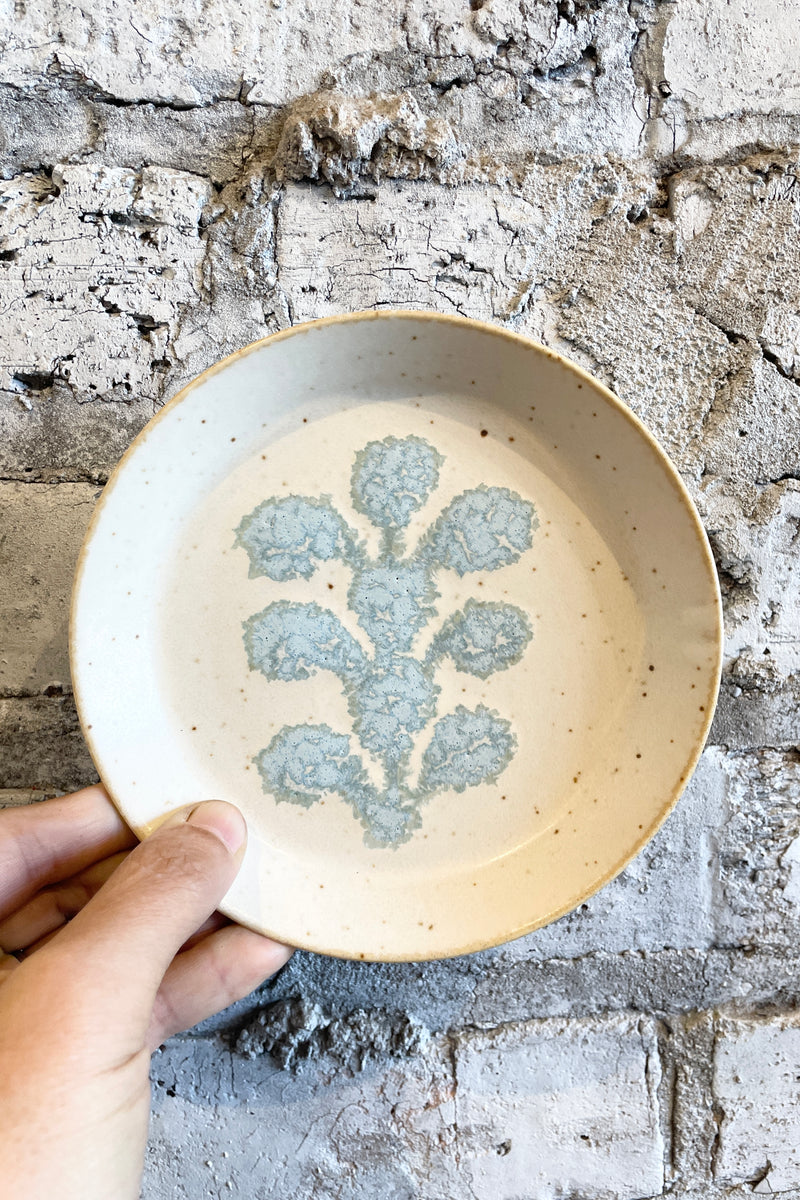 5.5" Ivory Blue Crest Plate being held in hand against a white brick wall. 