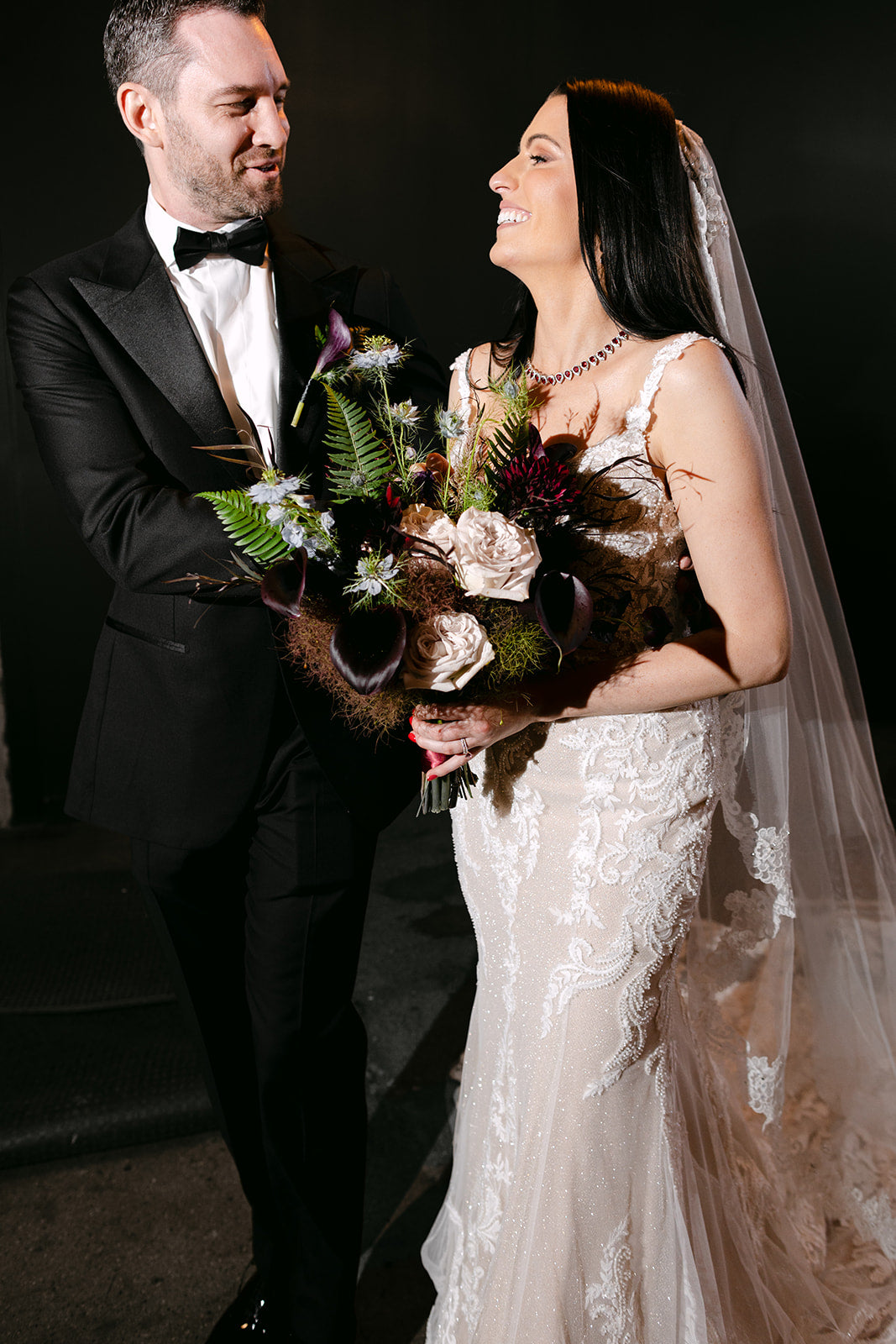 Groom in a tuxedo and Bride  in a wedding dress holding a dark and sexy bridal bouquet designed by Sprout Home against a dark background ©Devae Photography