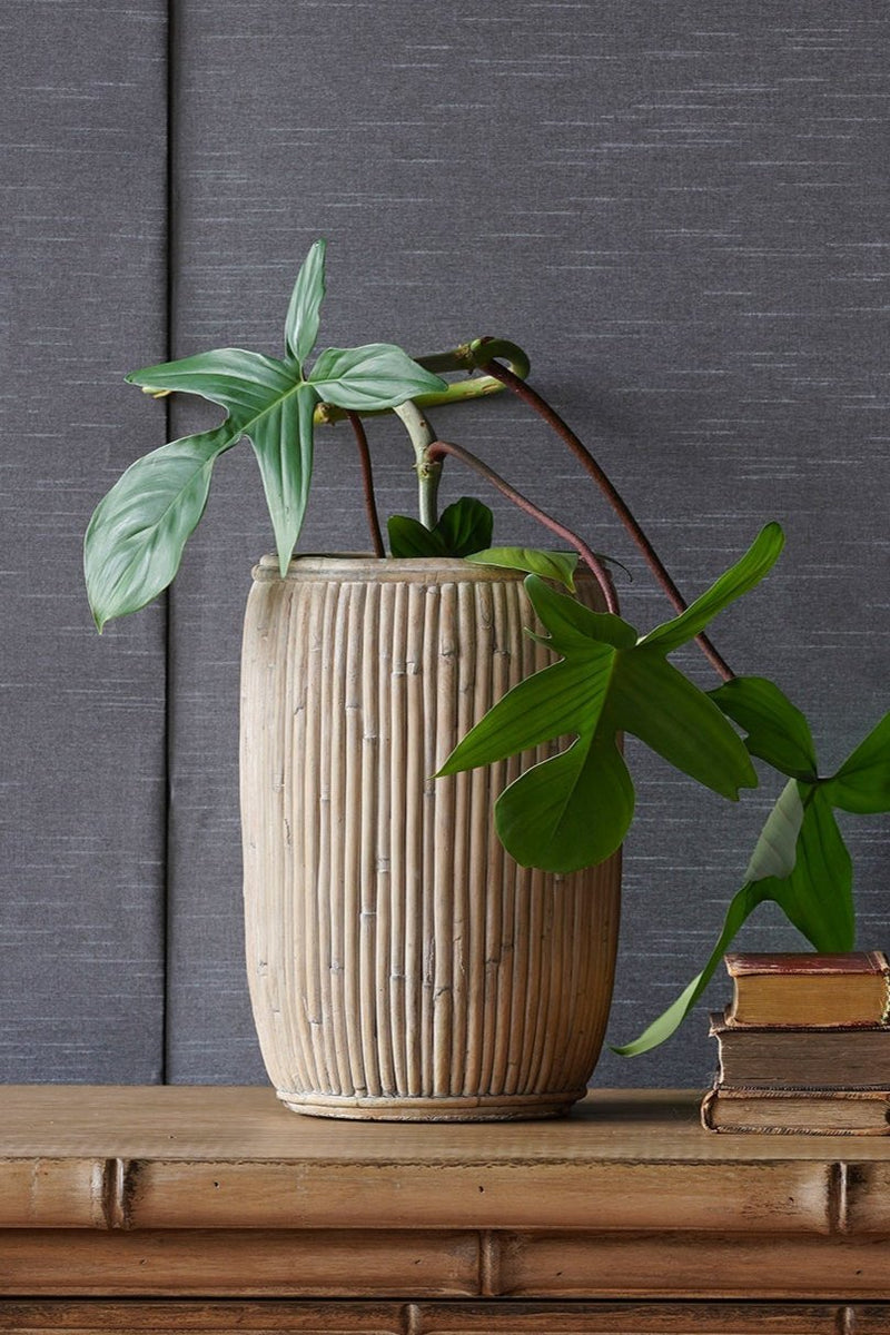 Large tall concrete faux rattan planter with a plant inside against a gray fabric wall and on top of a wood dresser