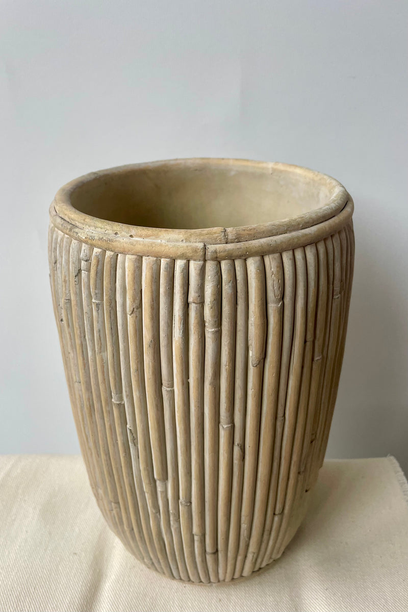 Medium tall concrete faux rattan planter against a white wall on a white tablecloth shown slightly from above.