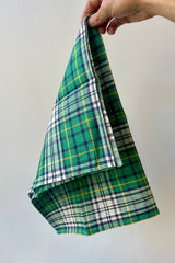 Phot of a hand holding a Helen Green plaid linen towel from the corner in front of a white wall. The Plaid is mostly green with dark blue white and yellow.