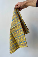 Photo of a hand holding an amber yellow linen towel in front of a white wall, pinched from the corner. The plaid is mostly yellow with taupe and light blue.