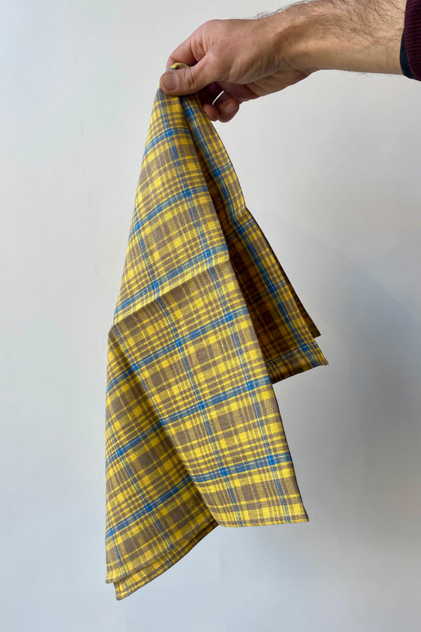 Photo of a hand holding an amber yellow linen towel in front of a white wall, pinched from the corner. The plaid is mostly yellow with taupe and light blue.