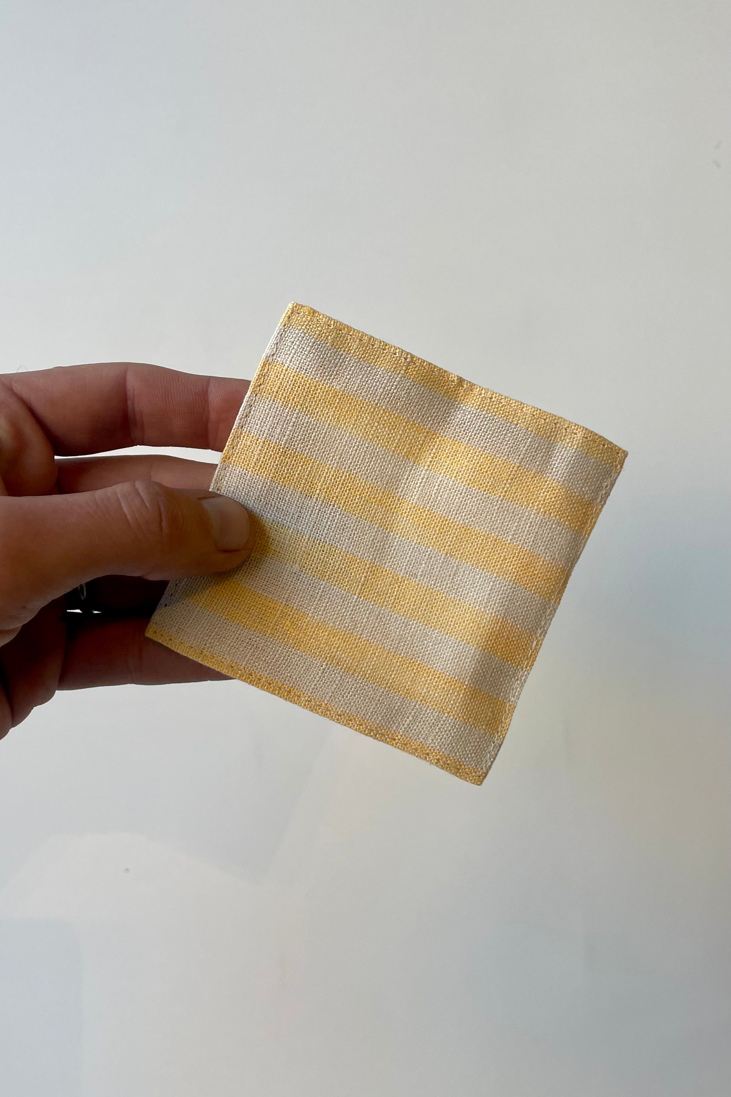 Photo of hand holding a yellow and beige striped square linen coaster in front of a white wall.