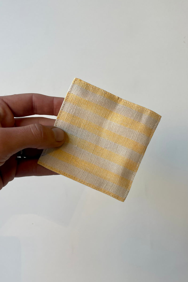 Photo of hand holding a yellow and beige striped square linen coaster in front of a white wall.