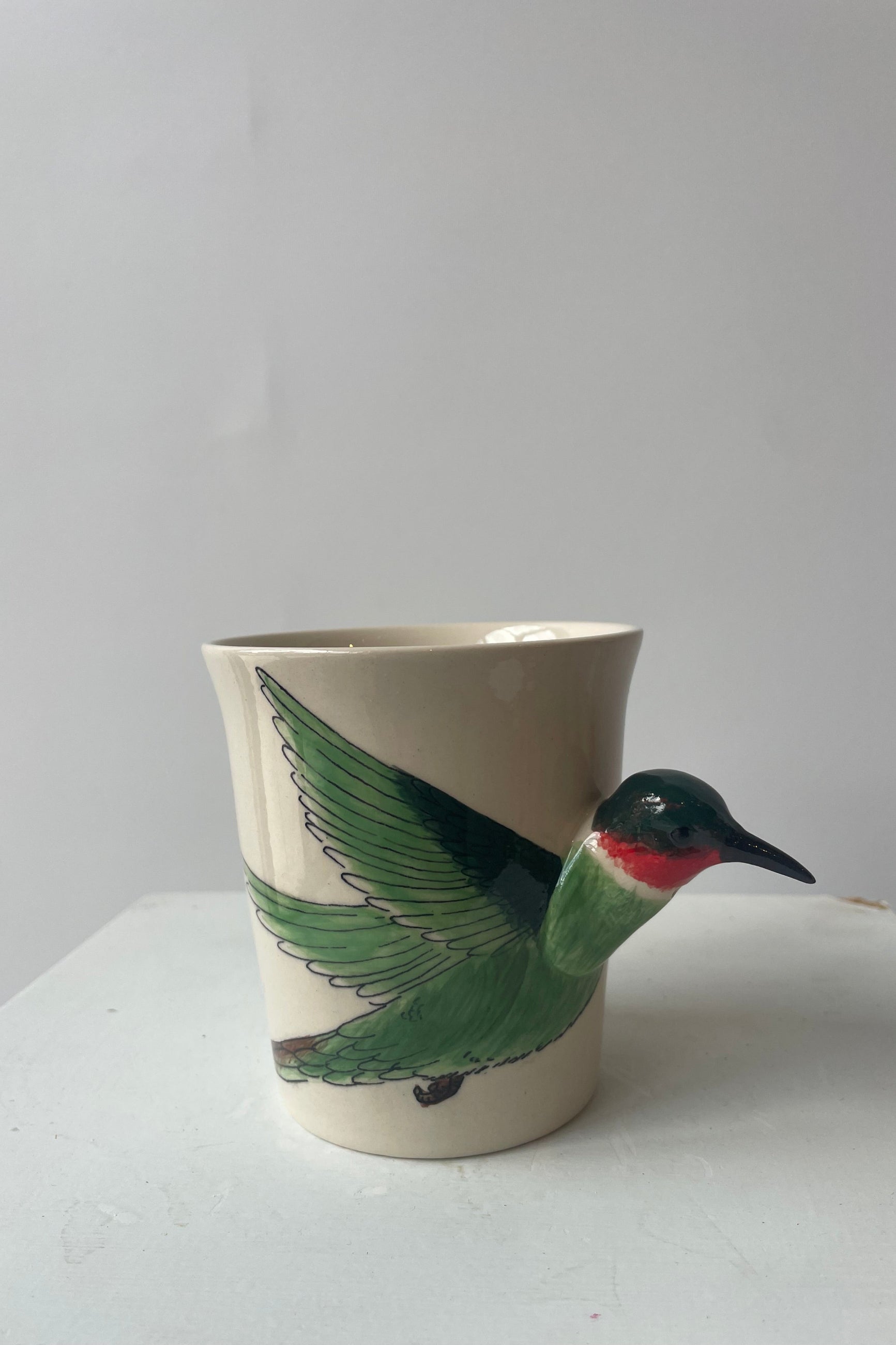 A white mug sits on a white table in a white room at Sprout Home. A green and ruby throated hummingbird shapes the side and handle of the mug. The mug sits upright.
