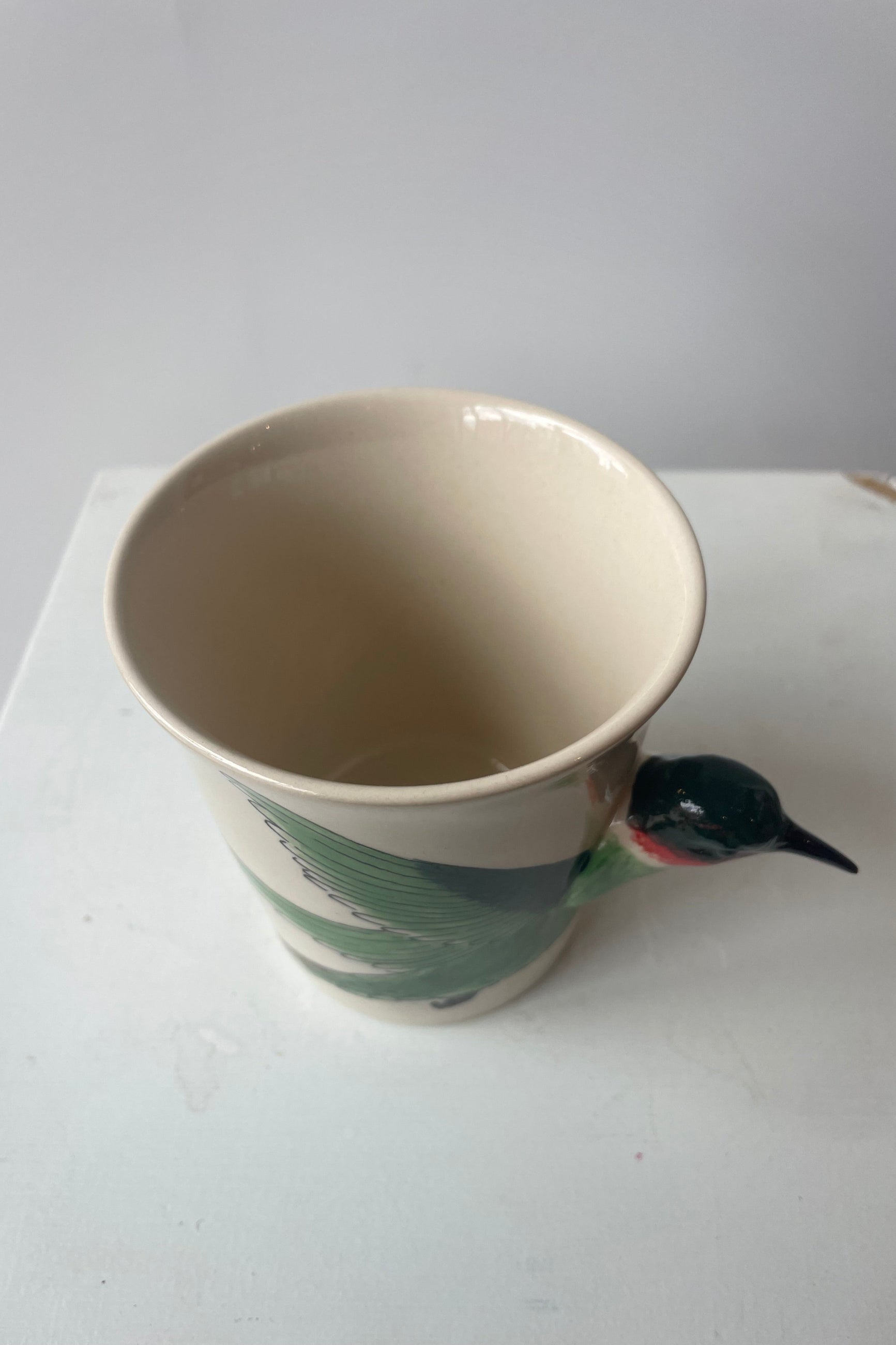 A white mug sits on a white table in a white room at Sprout Home. A green and ruby throated hummingbird shapes the side and handle of the mug.  The view is looking down into the mug.