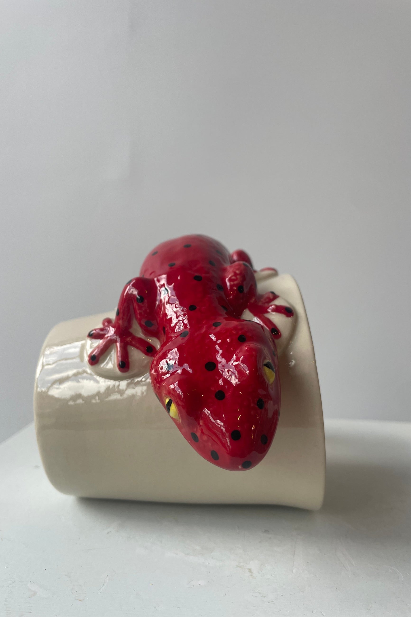 Photo of a white mug on a white table in a white room. A red salamander shapes the body and handle of the mug. The mug sits on its side at Sprout Home.