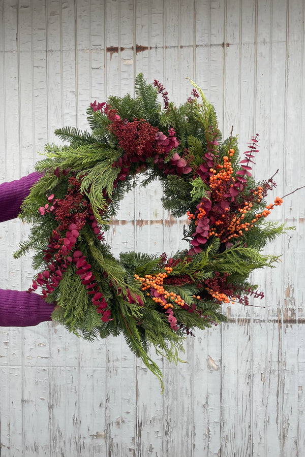 Photo of someone holding an evergreen wreath with burgundy and orange details of leaves and berries against a gray wall. The person is out of the frame with arms extended holding the wreath.