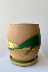 A round plant pot on a saucer. The pot is made of beige clay with a darker speckle pattern. There is a semi-gloss glaze of two shades of green dripped across the planter. The pot is photographed in a white room.