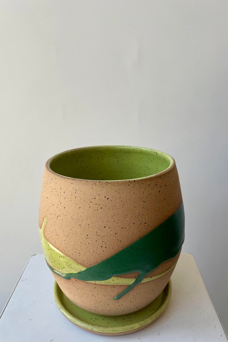 A round shaped plant pot on a saucer. The pot is made of beige clay with a darker speckle pattern. There is a semi-gloss glaze of two shades of green dripped across the planter. The pot is photographed in a white room. The angle of the photo shows the interior green glaze.