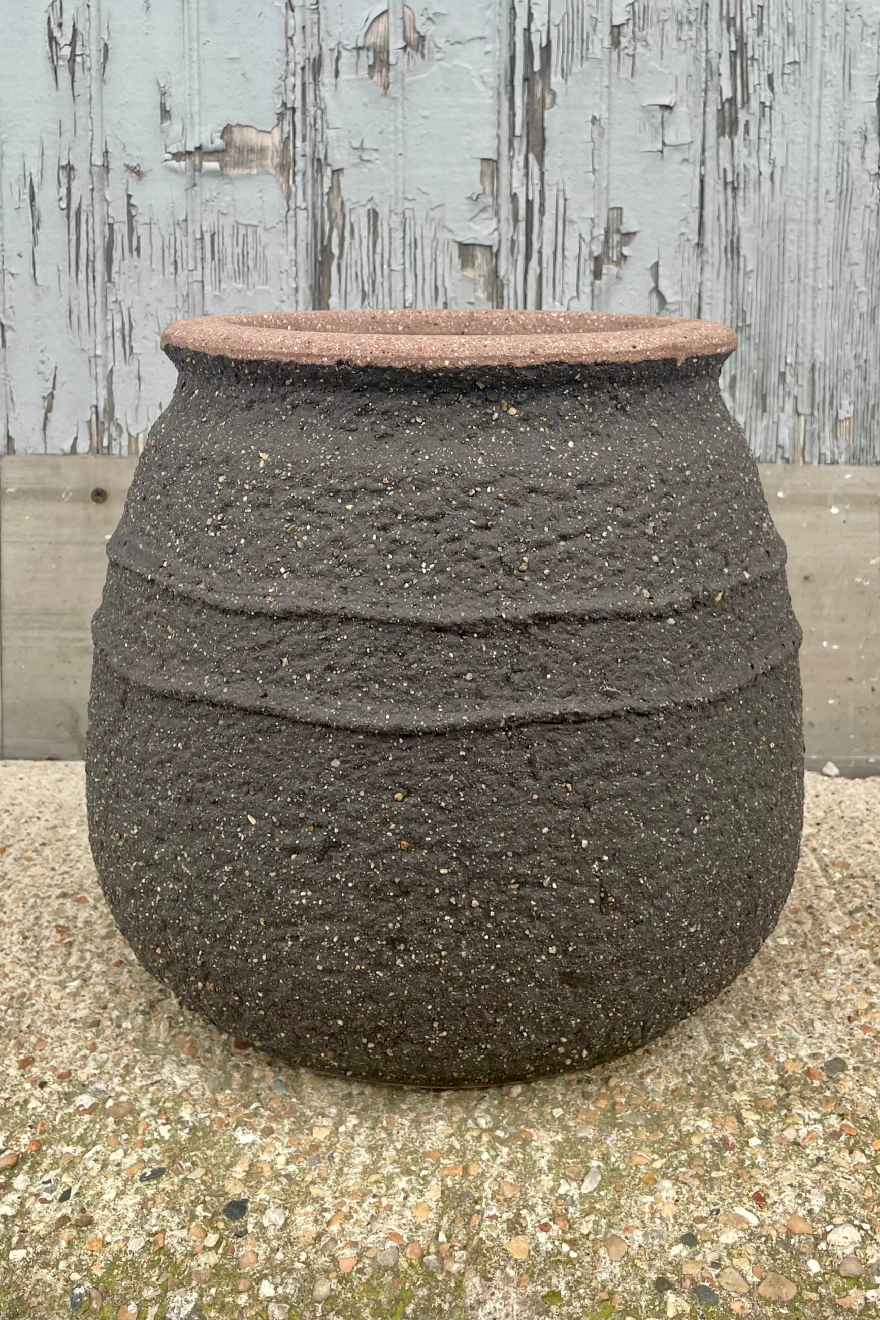A brown clay pot with a central textural band in front of a wooden wall. Show from the front ©Sprout Home
