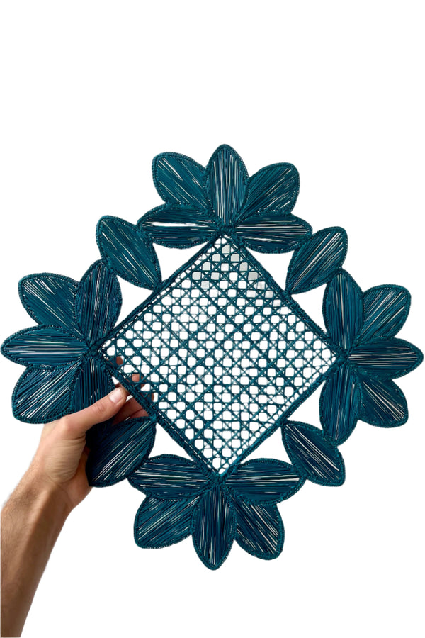 Photo of a Blue Square Leaves placemat of blue palm fiber and wire. It is held by a hand in front of a white background.