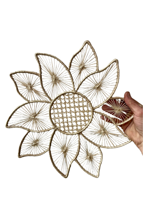 Photo of a hand holding a palm fiber and wire placemat. The design is Big Flower Araca of beige fiber and is shown against a white background.