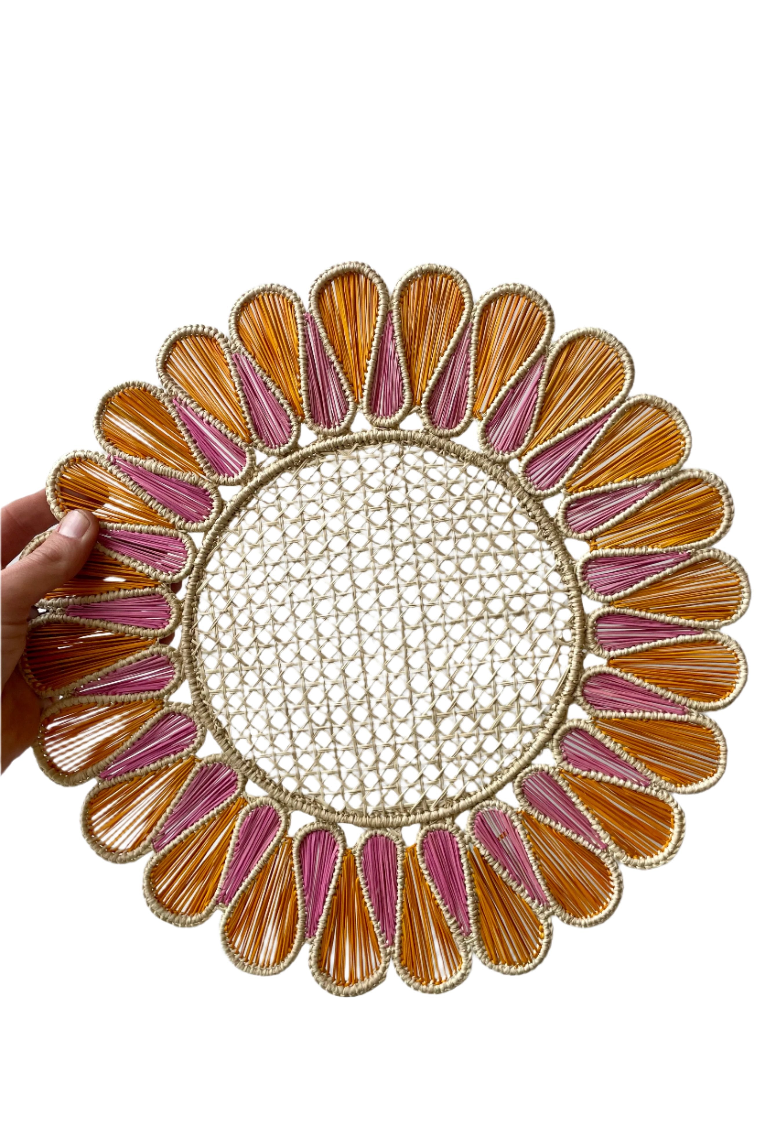 Photo of a Drops Placemat of Tangerine and Guava dyed palm fiber around wire. A hand is holding the placemat in front of a white background.