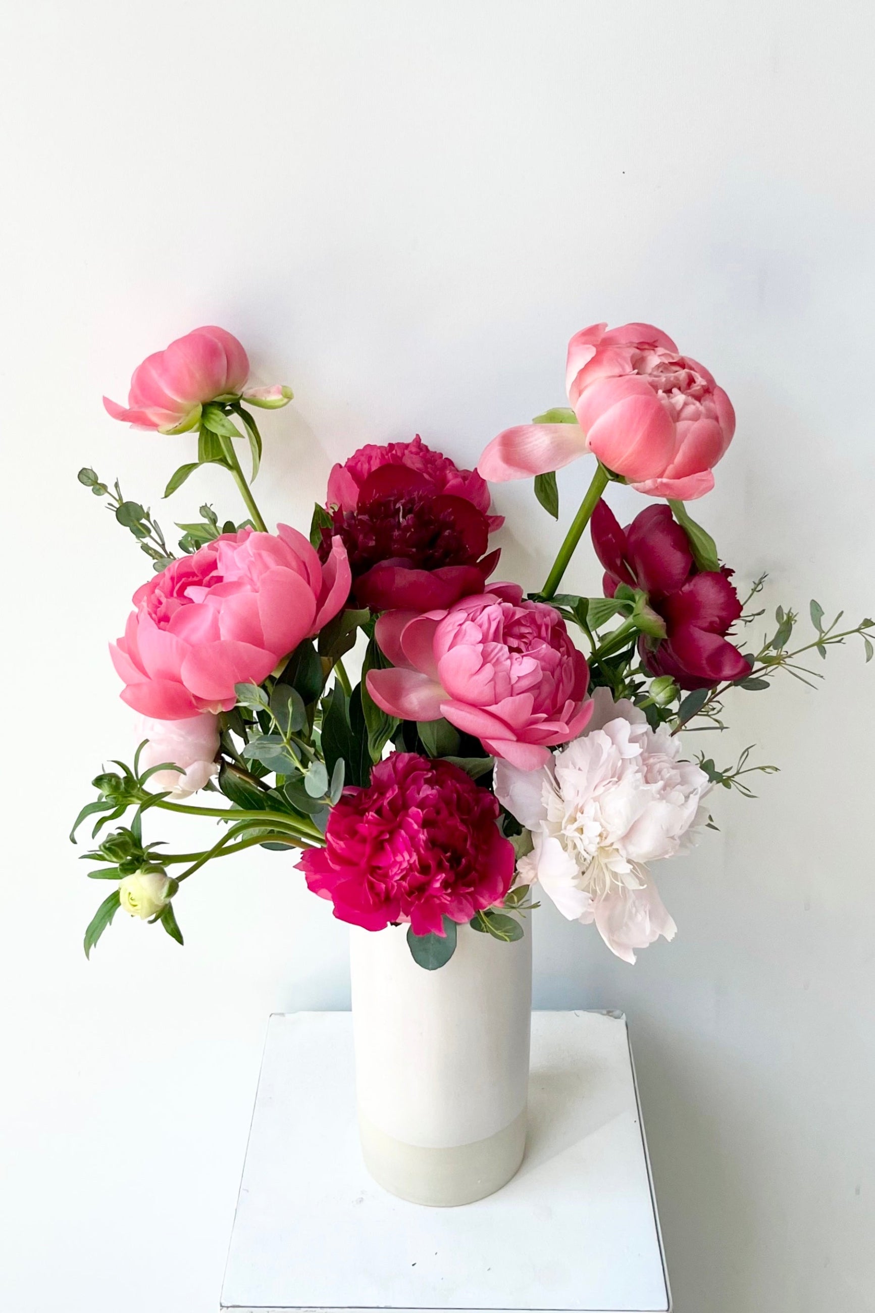 Bouquet of pink, red, and white Peony flowers in a white vase on a white surface with a light gray background ©Sprout Home #size_$130