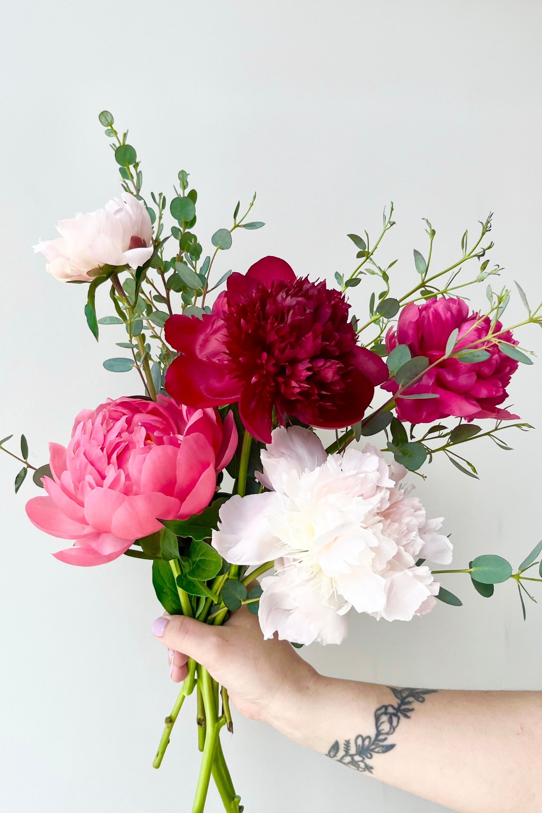 Bouquet of pink, red, and white Peony flowers held by a hand against a light gray background ©Sprout Home #size_$75