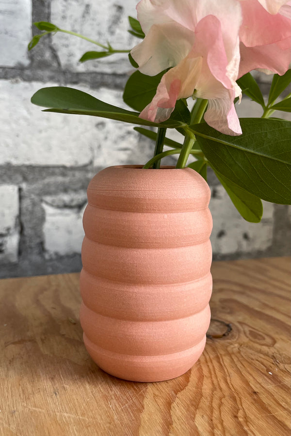 Peite Pastel Peach Ribbed vase on a wood surface in front of white brick. 